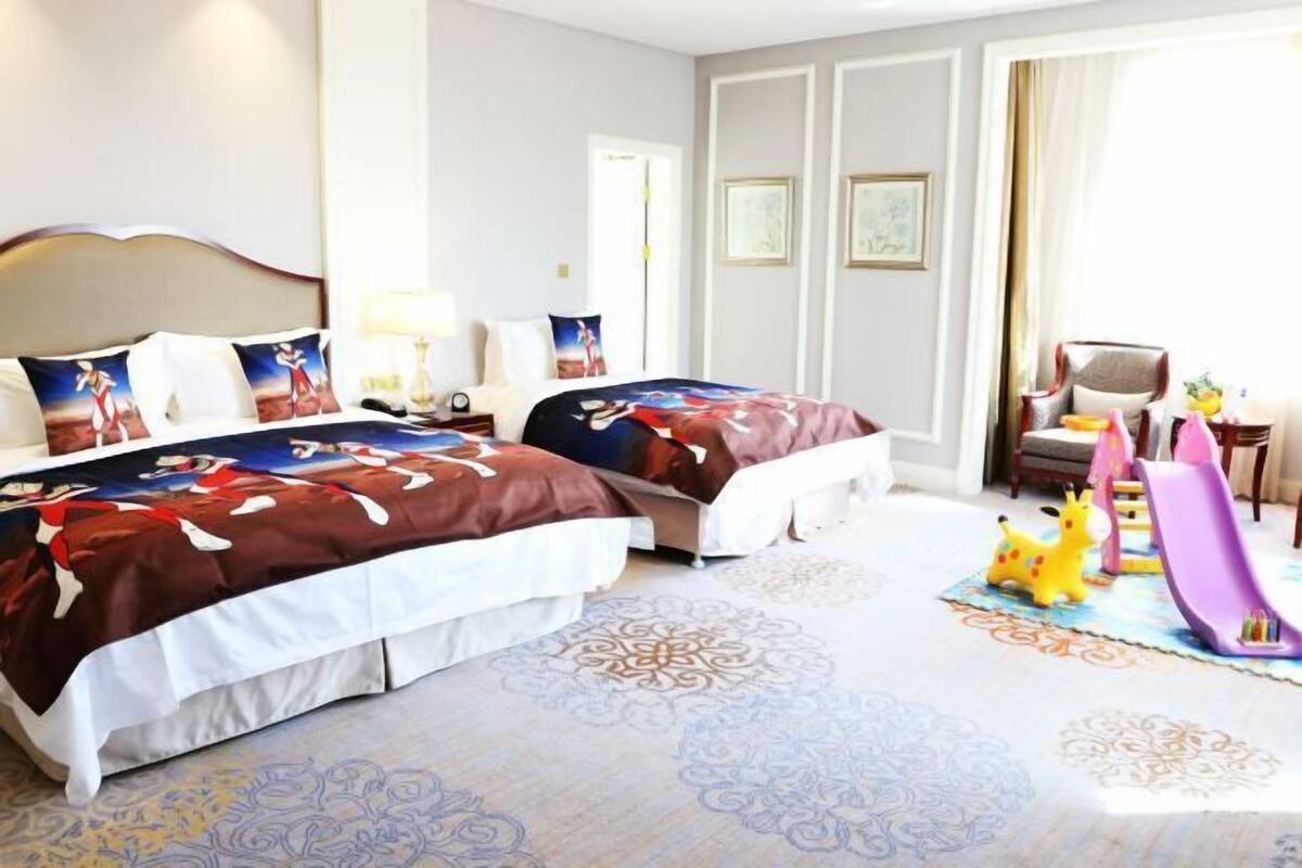 A hotel room with two beds, a desk, a chair, a desk lamp, a small table, a toy giraffe, a toy slide, and a patterned rug.