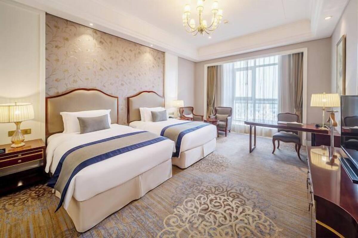 A hotel room with two beds, a desk, a chair, a TV, and a large window.