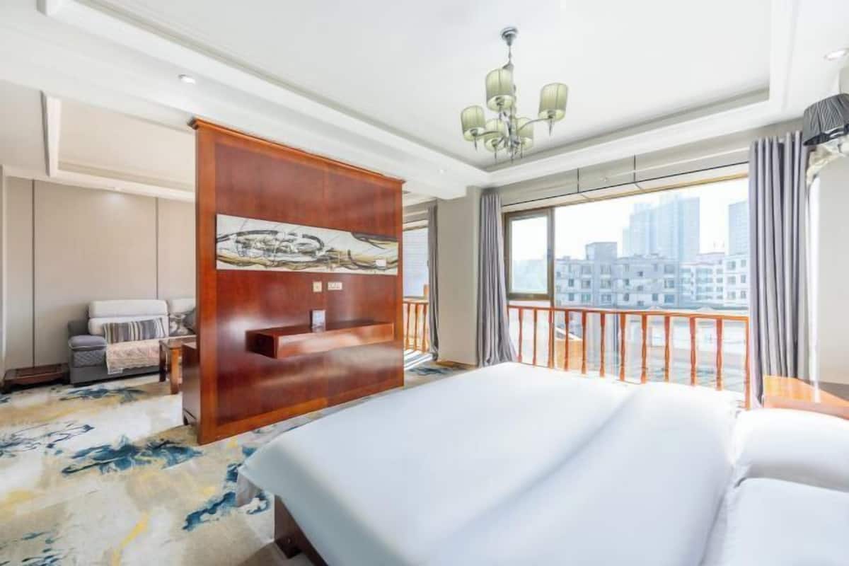 A hotel room with a large bed, a wooden partition with artwork, a balcony with a railing, and a city view.