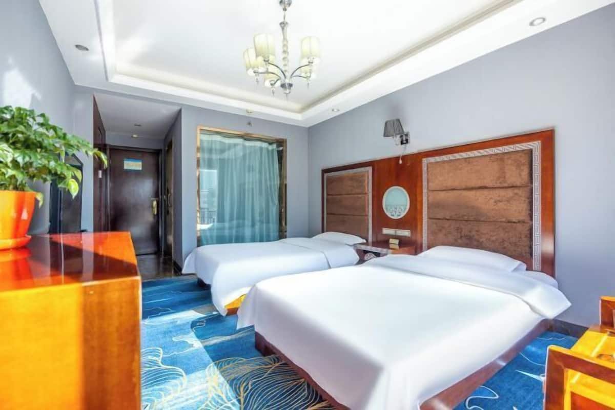 A hotel room with two beds, a chandelier, a large window with curtains, and a potted plant.