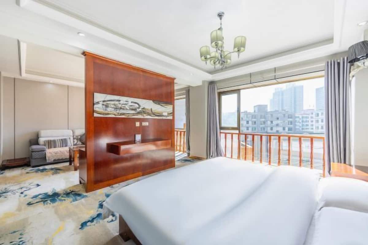 A hotel room with a large bed, a wooden partition with artwork, a balcony with a railing, and a city view.