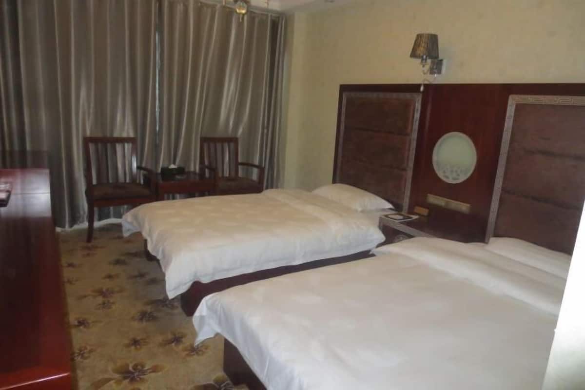 A hotel room with two beds, a wooden headboard, a small table, a chair, and a curtain.