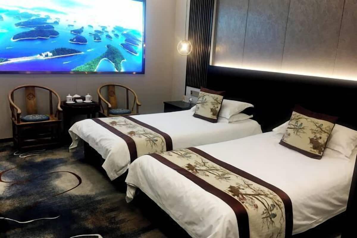 Habitación de hotel con dos camas, un televisor mostrando una vista panorámica, sillas de madera y una mesita con un calentador de agua.