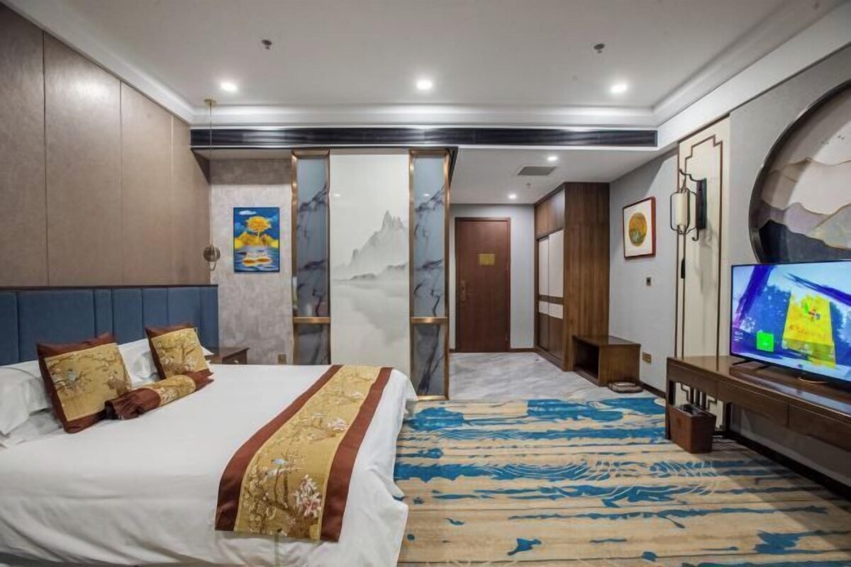 A hotel room with a large bed, a television, a desk, and a view of a mountain through a glass partition.