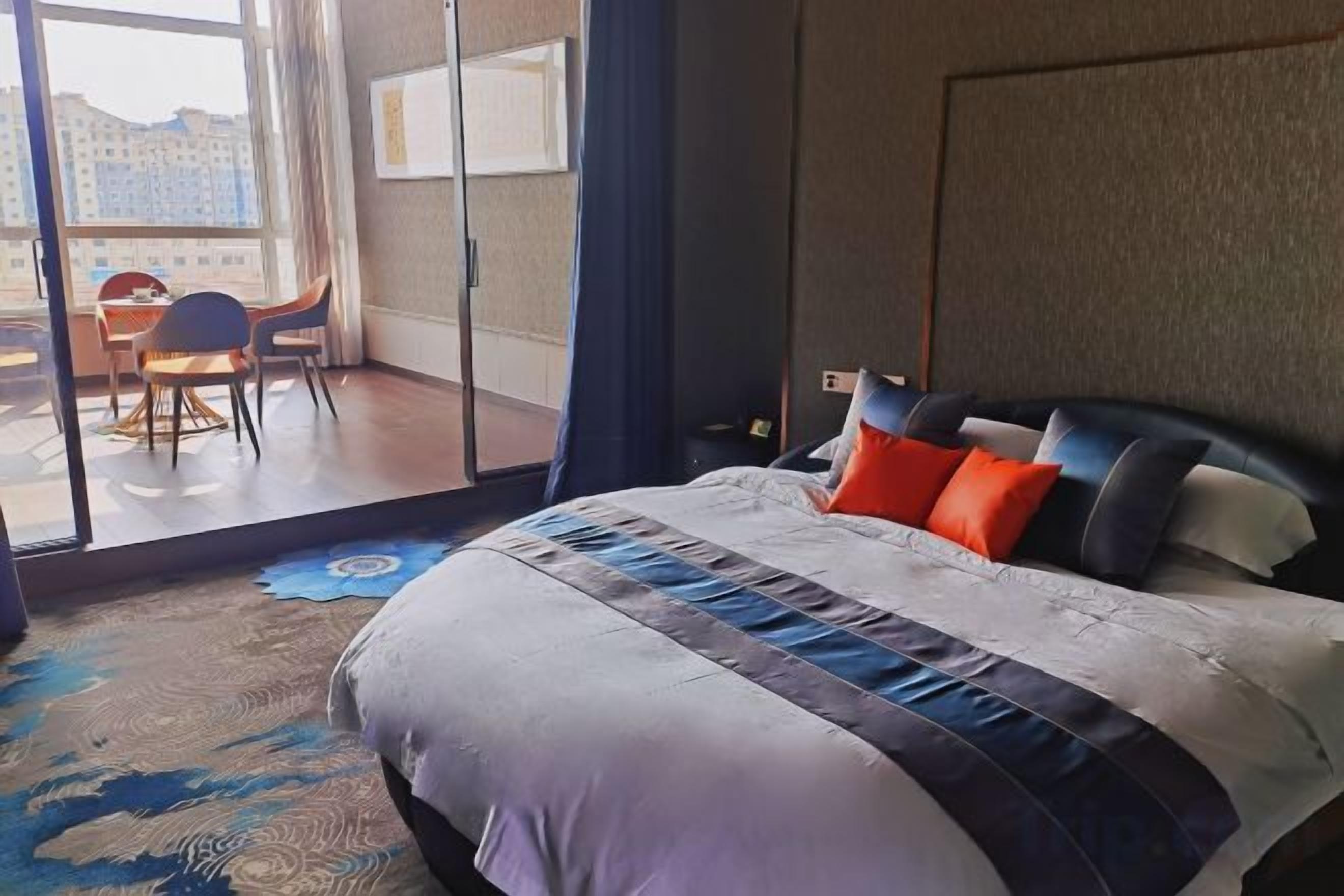 A hotel room with a large bed, a view of the city through a sliding door, and a balcony with a table and chairs.