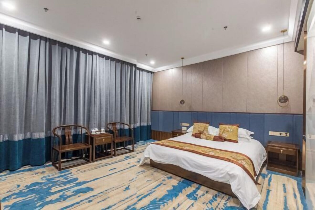 A hotel room with a large bed, two wooden chairs, a nightstand, and a blue and beige patterned carpet.