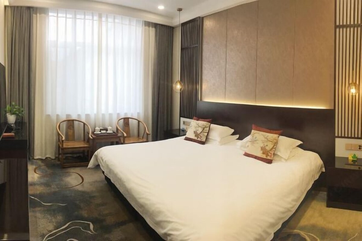 A hotel room with a large bed, a desk with a chair, a bedside table, and a large window with curtains.