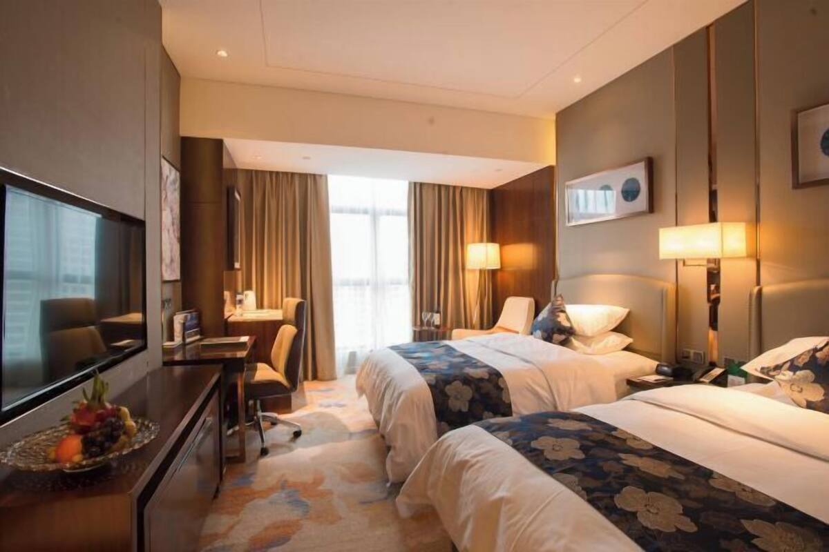 A hotel room with two beds, a desk, a TV, and a dining table with fruit.