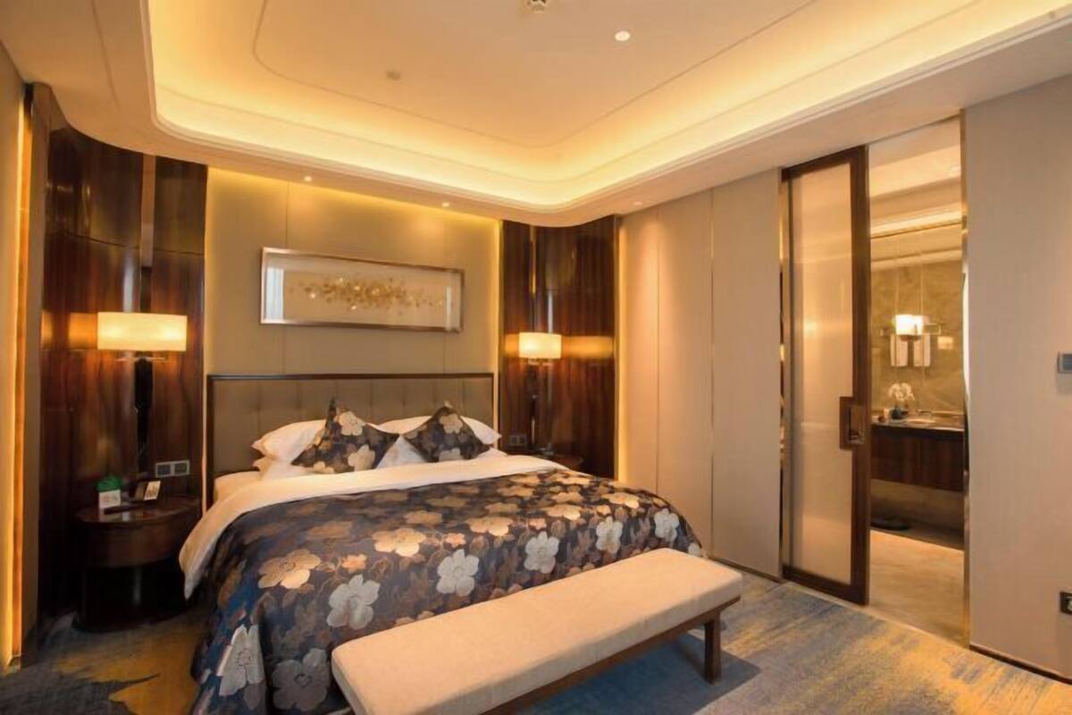 A hotel room with a large bed, bedside lamps, a bench, a mirror, and a bathroom visible through an open door.