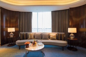 A modern hotel room with a curved sofa, two side tables with lamps, a coffee table with fruit and a bottle, and a large window with curtains. - Phoenix Hotel (Wenshan City)