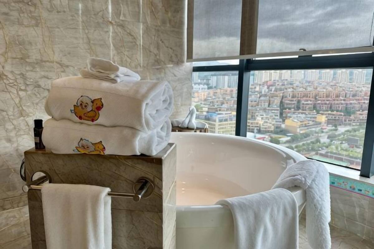 A bathroom with a bathtub, towels, and a city view through large windows.