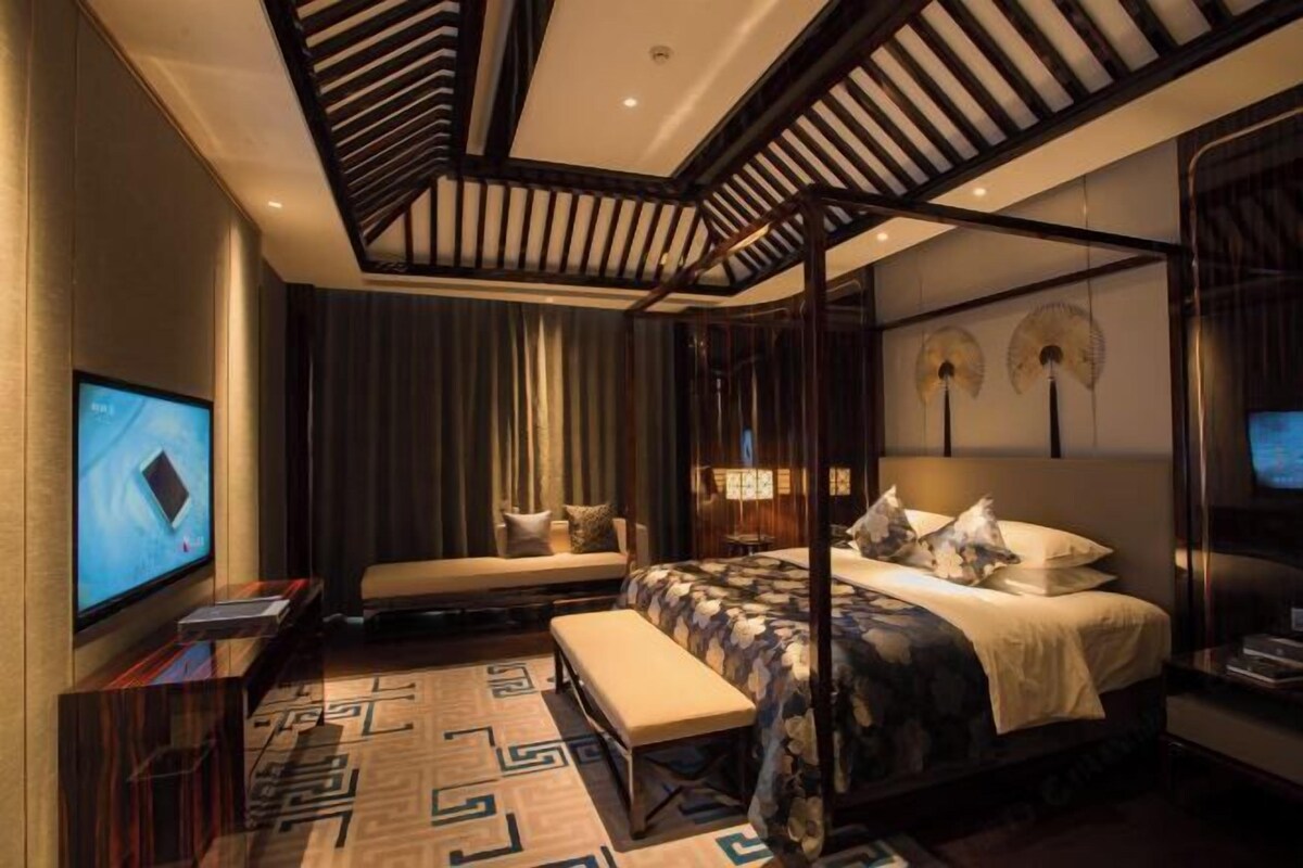 A modern hotel room with a large bed, a bench, a TV, and a unique ceiling design.