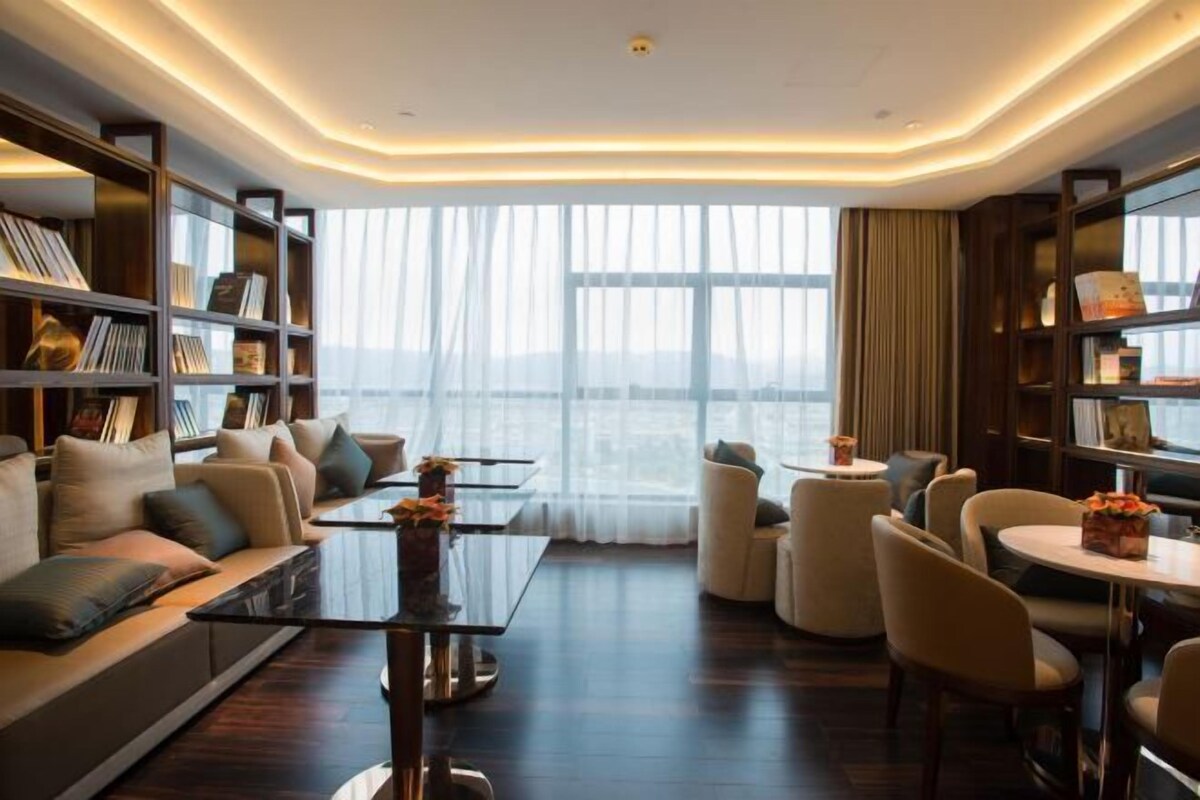 A modern hotel room with a large window, a sofa, a coffee table, and a bookshelf.