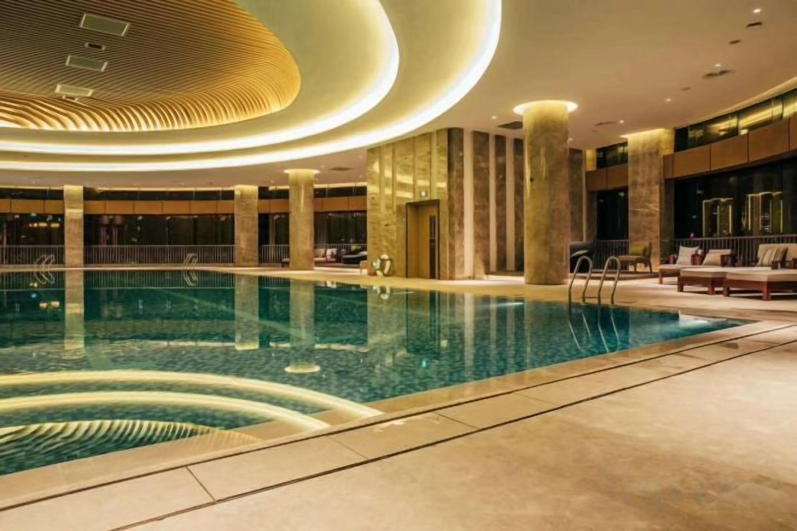 A large indoor swimming pool with a modern design, surrounded by a lounge area with seating and a poolside bar.