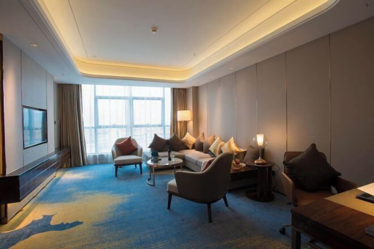 A hotel room with a large window, a sofa, a chair, a coffee table, and a desk with a lamp.