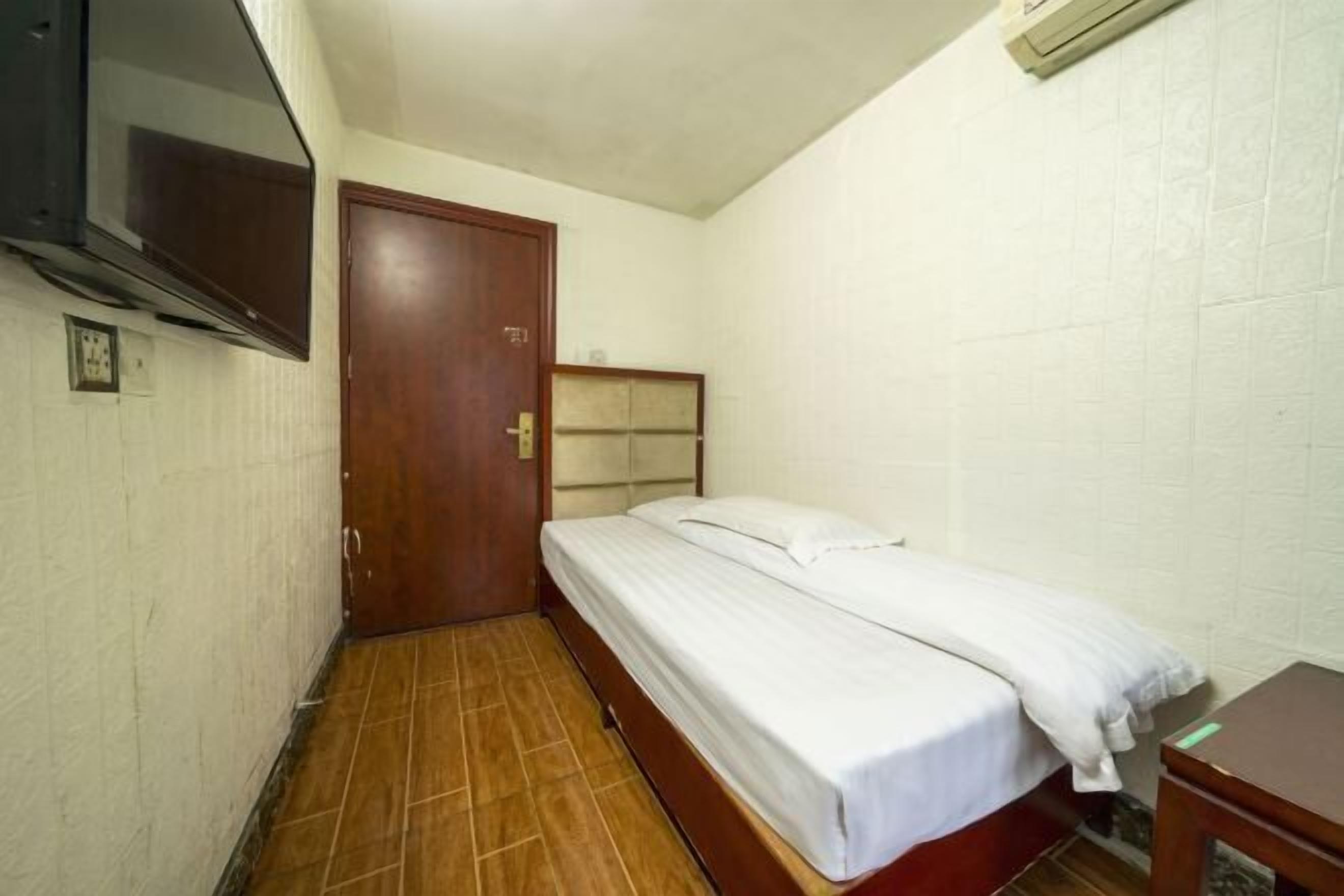 Une petite chambre avec un lit simple, une porte en bois, un téléviseur fixé au mur et un sol carrelé.