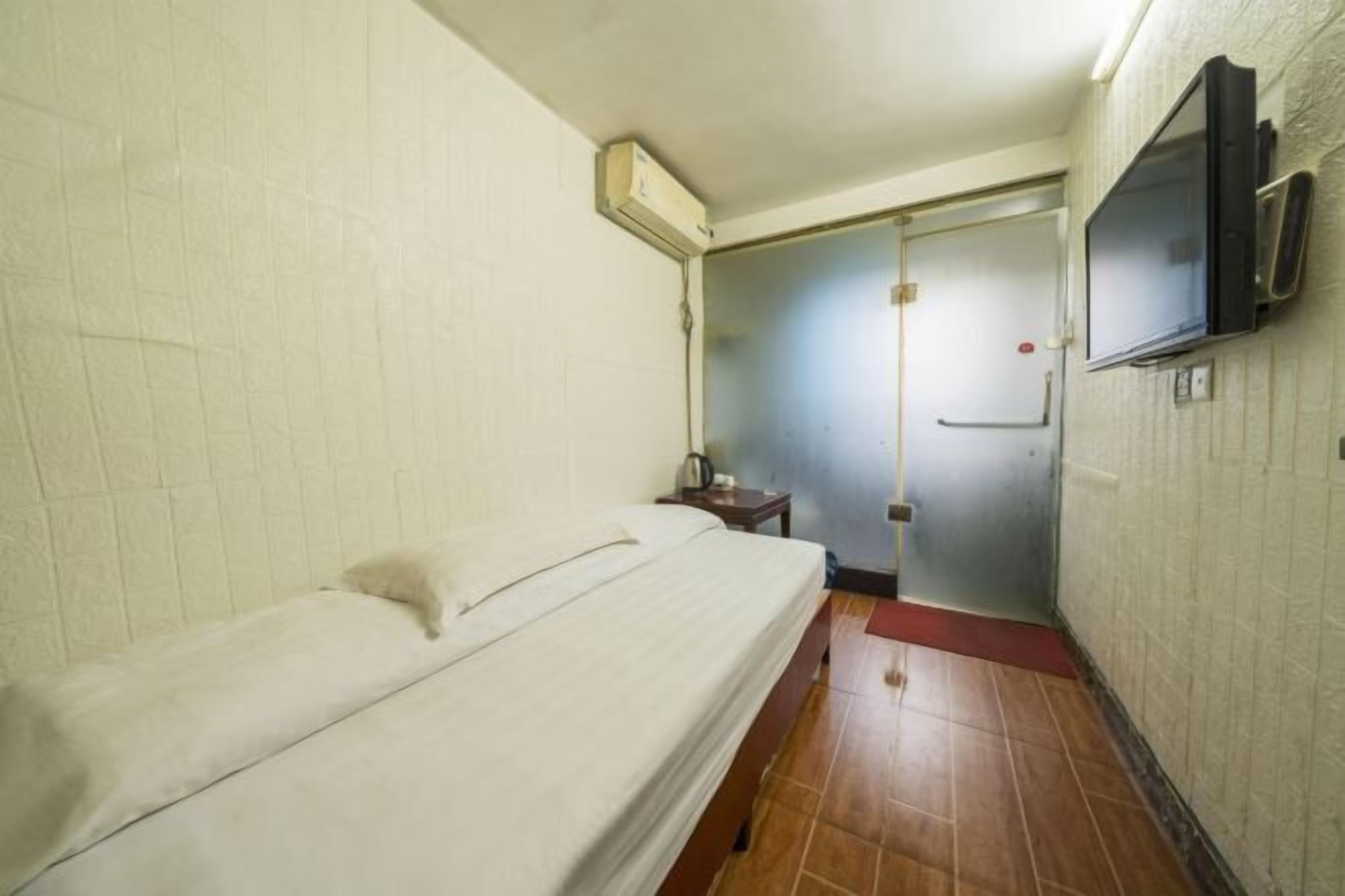 A small, compact room with a single bed, a television mounted on the wall, and a door leading to another room.