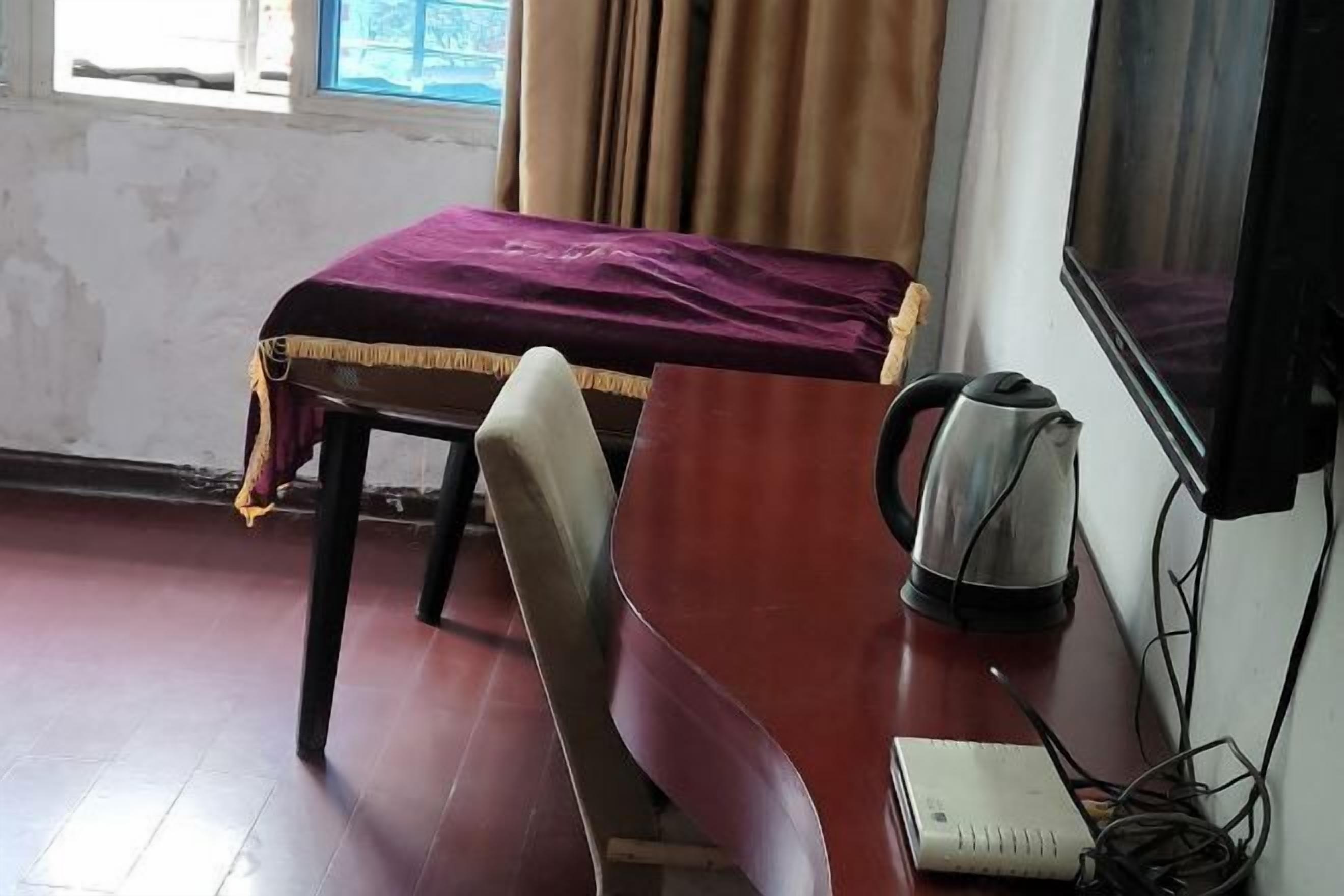 Une chambre avec un lit recouvert d’une couverture violette, un bureau en bois avec une bouilloire et un carnet.