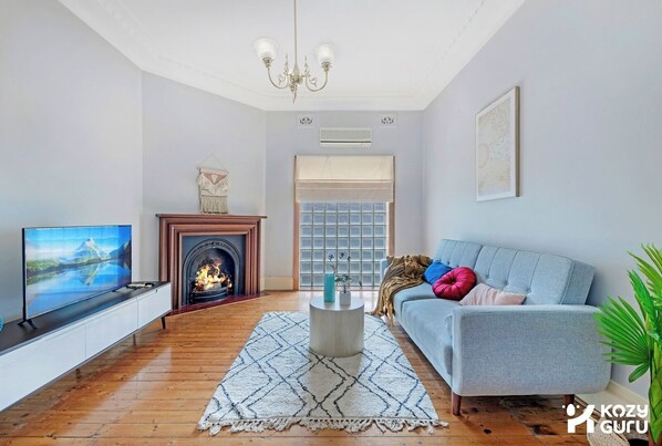 A living room with a fireplace, a sofa, a TV, and a large window. - KozyGuru | Campsie | Oldie-Goodie Look 4 Bed House (Campsie)