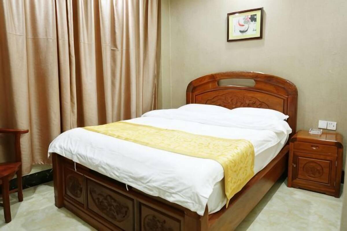 Una cama de madera con colcha blanca y amarilla, una mesita de noche, una silla y un cuadro en la pared.