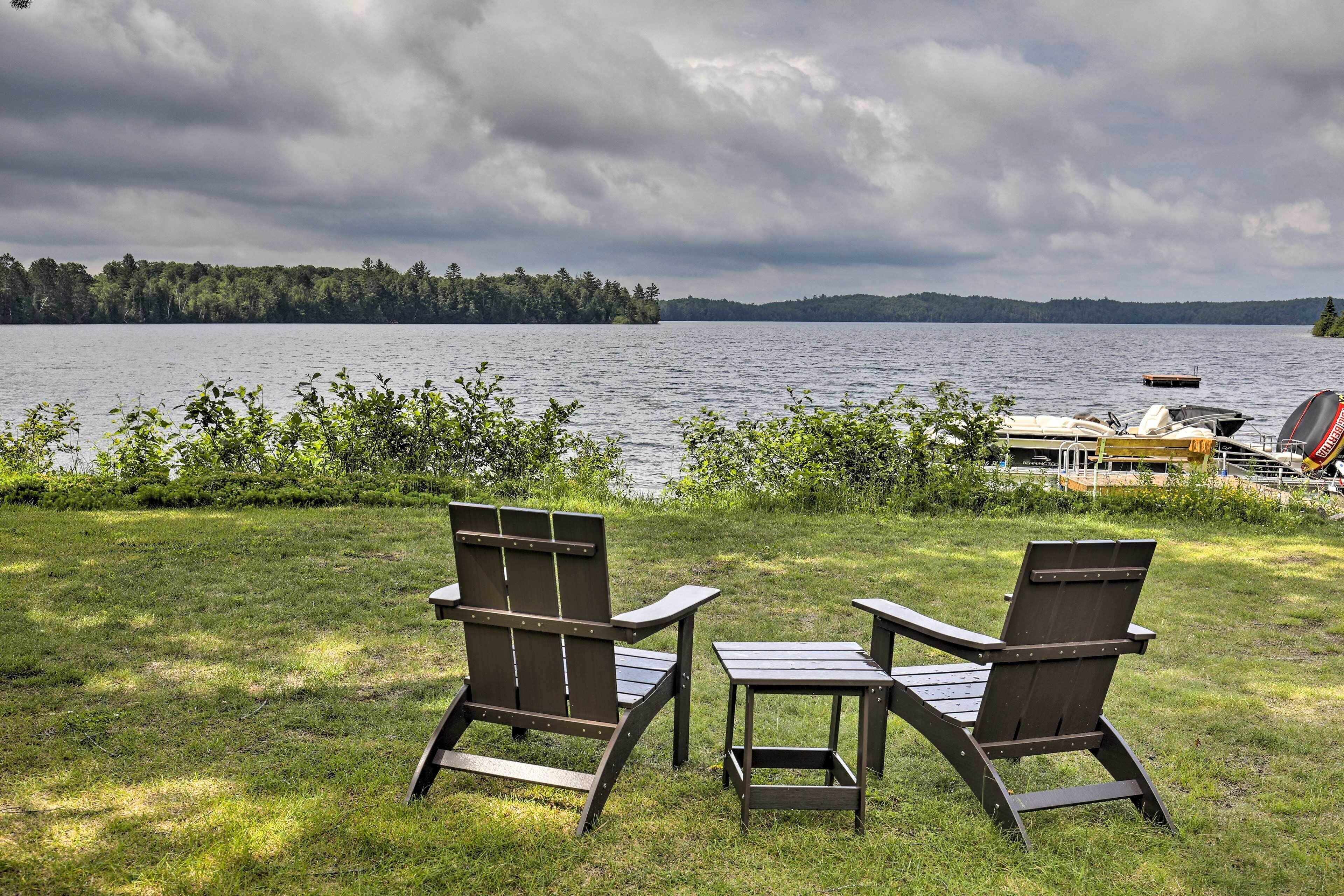Two Adirondack chairs and a small table on a grassy area overlooking a lake with boats and a dock in the background.