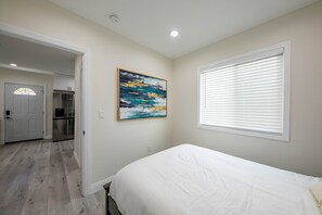 A bedroom with a bed, a painting, a window with blinds, a door, and a kitchen area.