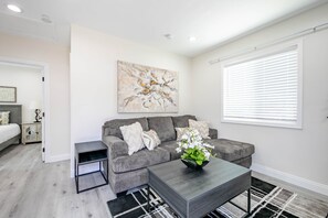 A modern living room with a grey sofa, a coffee table with a flower vase, a painting on the wall, and a window with blinds.