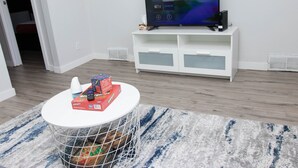Una sala moderna con un televisor de pantalla plana sobre un soporte blanco, una mesita pequeña con una cesta, y una alfombra azul y blanca.