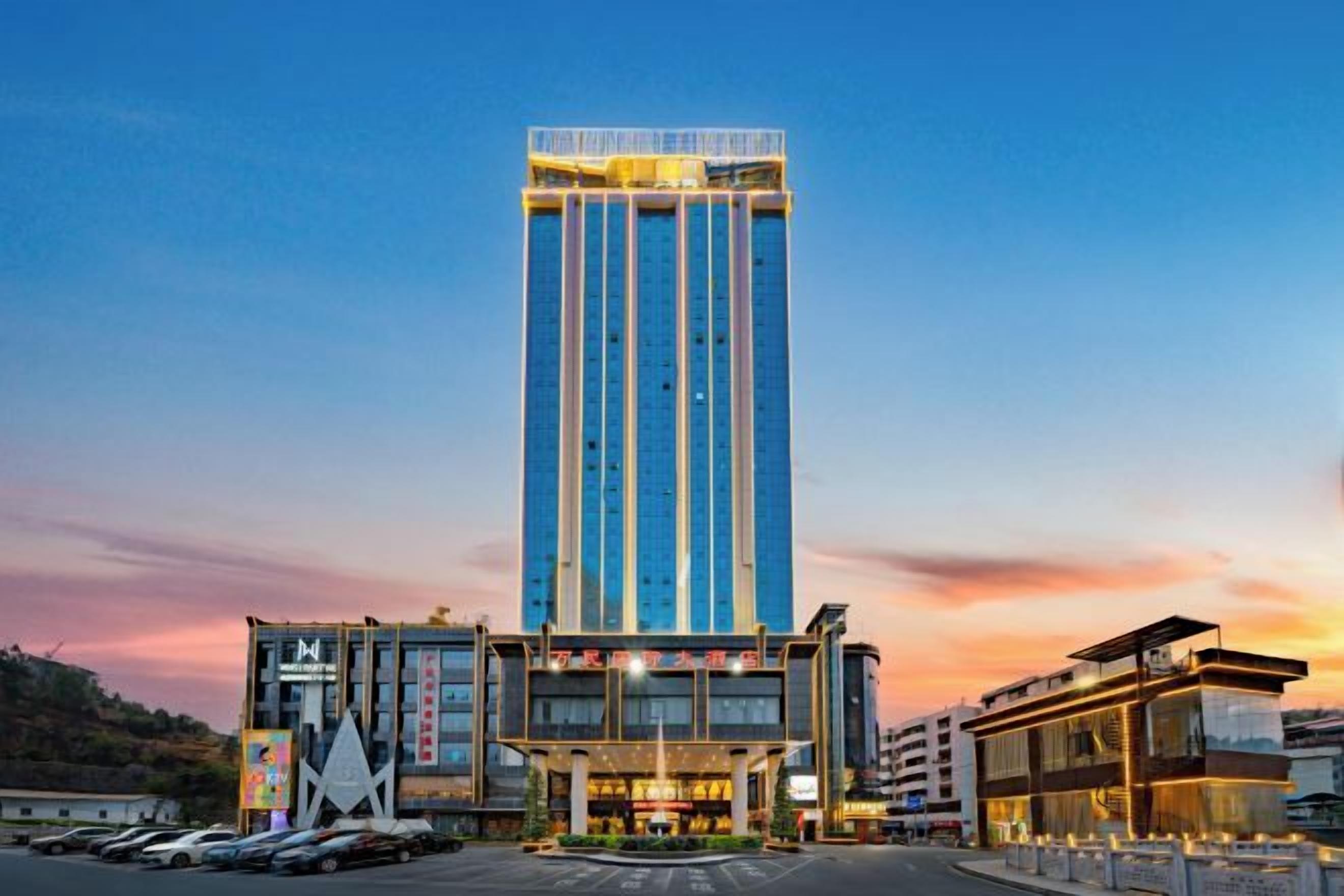A modern hotel with a tall central tower, a ground-level building with a glass facade, and a parking lot.