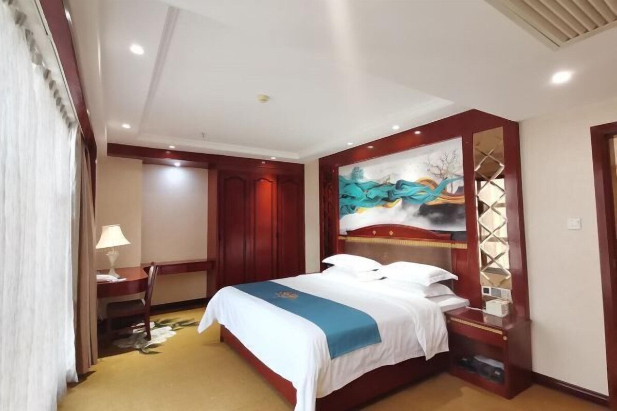 A hotel room with a bed, a desk, a chair, a lamp, and a painting on the wall.