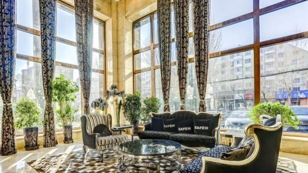 A spacious lobby with a checkered floor, a black sofa with 'SAFE' cushions, a round glass coffee table, and tall windows with patterned curtains. - Roman Garden Business Center (Mudanjiang)