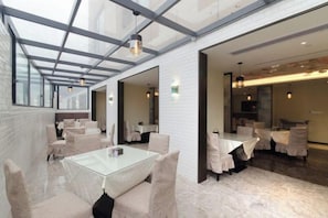 A modern dining area with glass tables and chairs, a high ceiling with a skylight, and a view of the adjacent room. - Huayi Hotel (Chongyang)