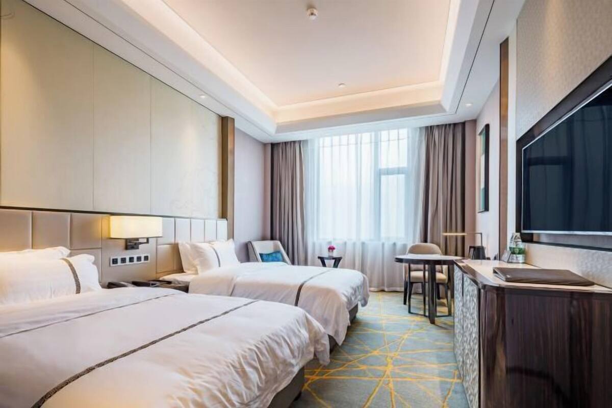 A hotel room with two beds, a desk, a TV, and a large window with curtains.