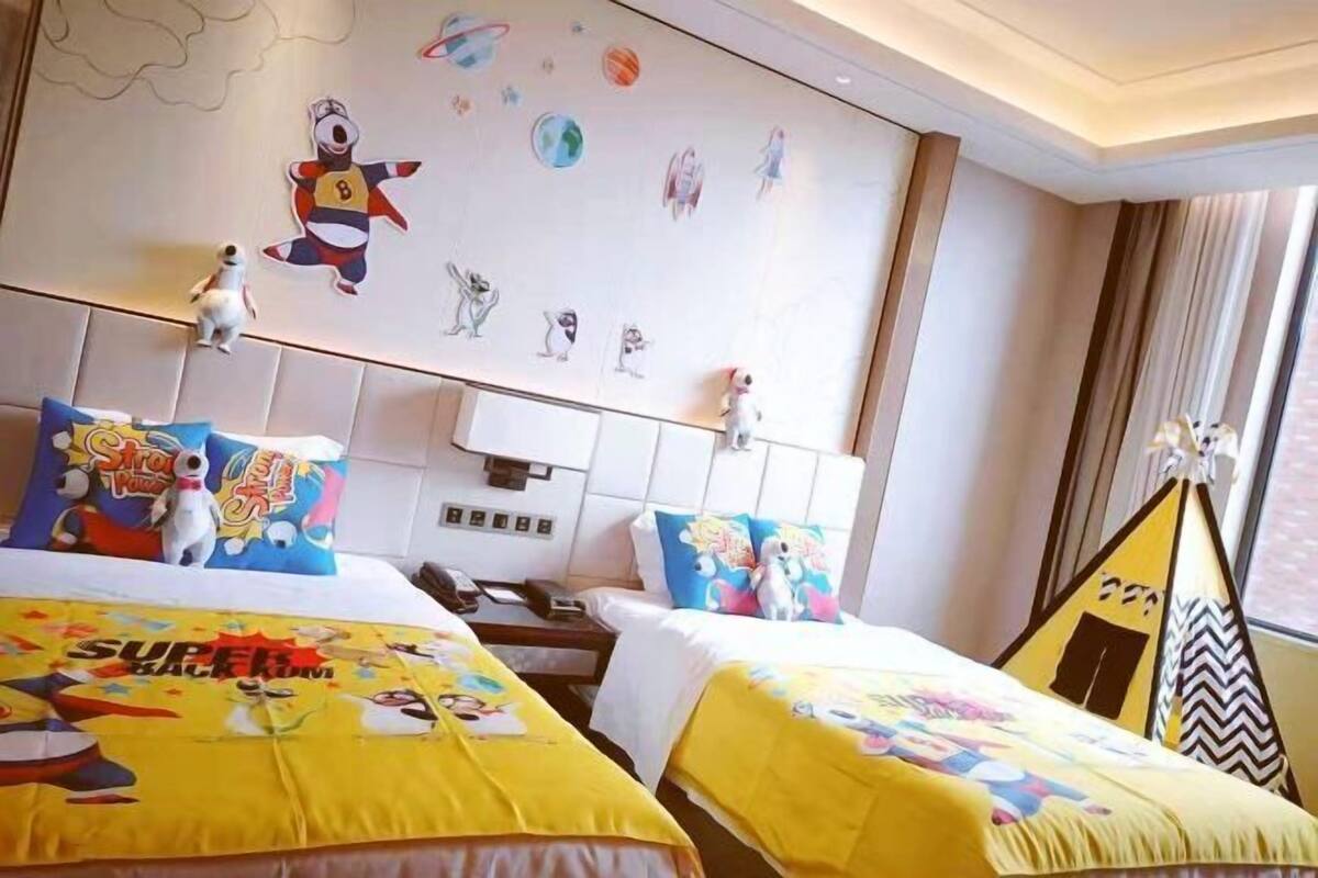 A hotel room with two beds, each with a yellow bedspread featuring cartoon characters. There is a wall with a space-themed mural, a small table with a phone, and a window with curtains.