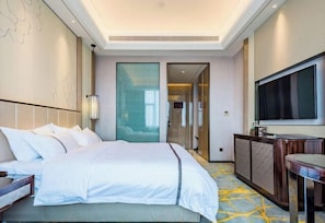 A hotel room with a bed, a desk, a TV, and a glass door leading to a hallway. - Huayi Hotel (Chongyang)