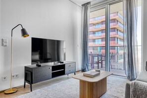 A modern hotel room with a flat-screen TV, a wooden side table, a desk, a floor lamp, and a balcony with a view of a multi-story building. - Classic Landstraße 1BR w/ Pool & DM, nr U3 Erdberg, by Blueground (Vienna)