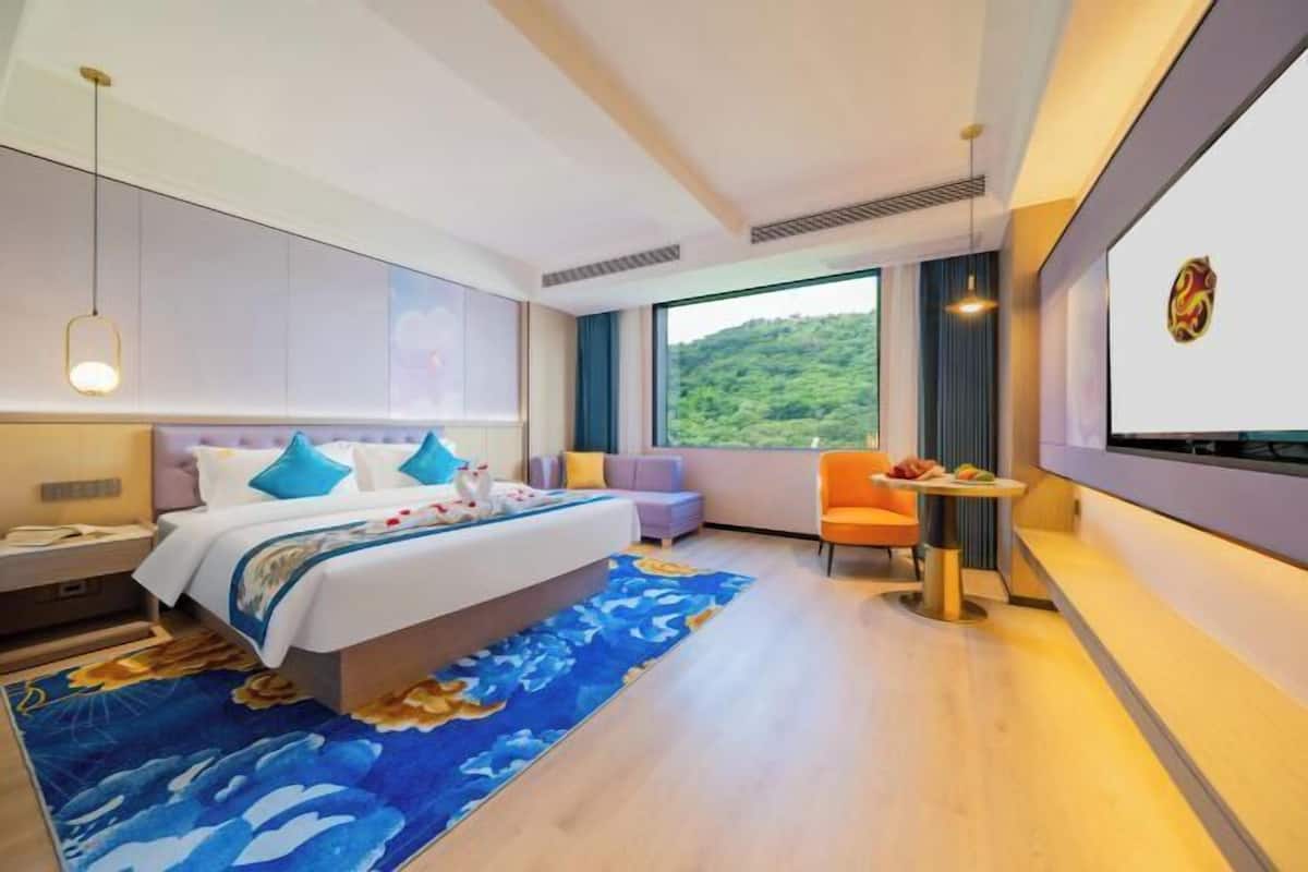 A modern hotel room with a bed, a small table, a chair, and a large window offering a view of greenery.