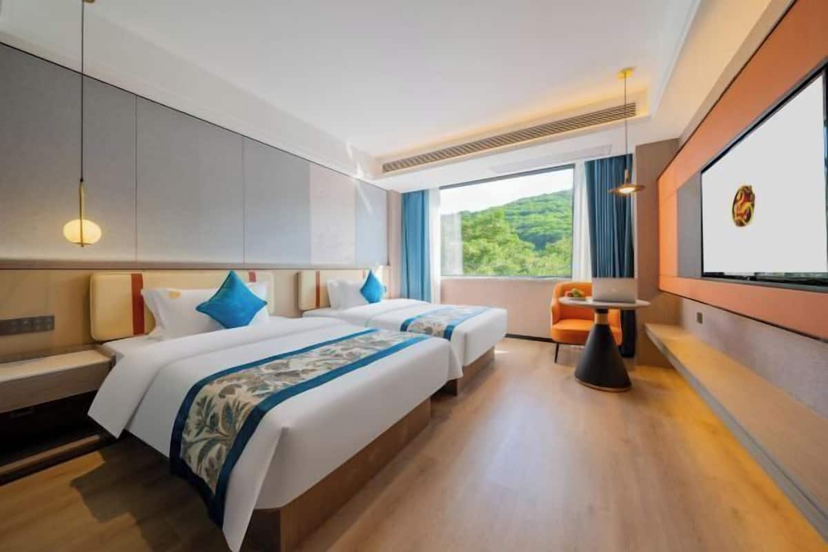 A hotel room with two beds, a desk, a chair, and a large window with a view of greenery.