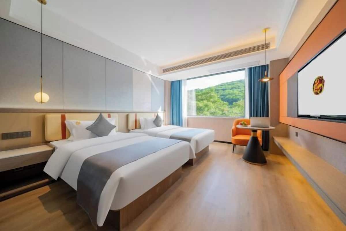 A hotel room with two beds, a desk, a chair, and a large window with a view of greenery.