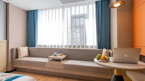 Una habitación de hotel moderna con un sofá cama, una mesita con una laptop y frutas, y un ventanal con cortinas.