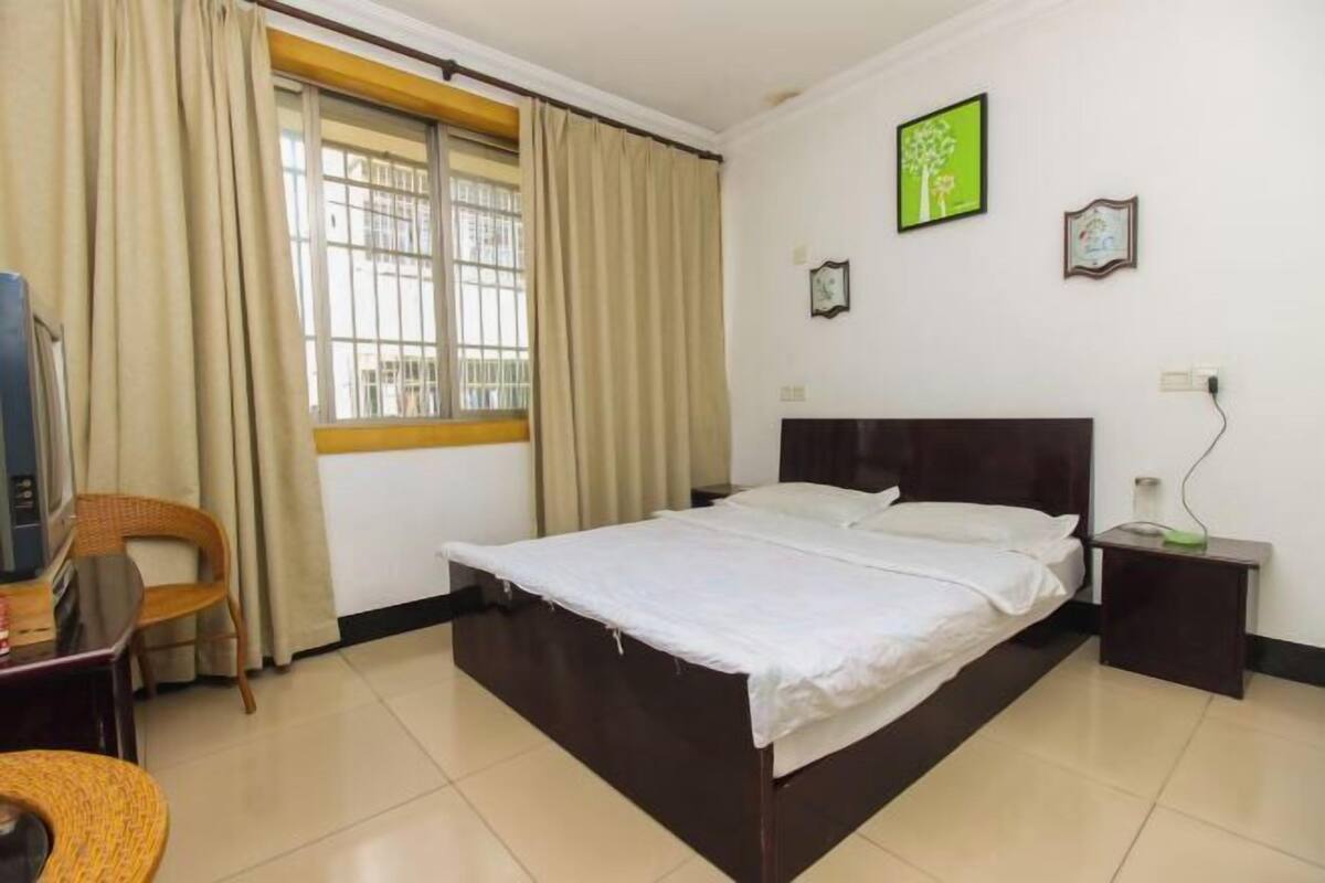 A bedroom with a bed, a chair, a desk, a window with curtains, and a wall with framed pictures.