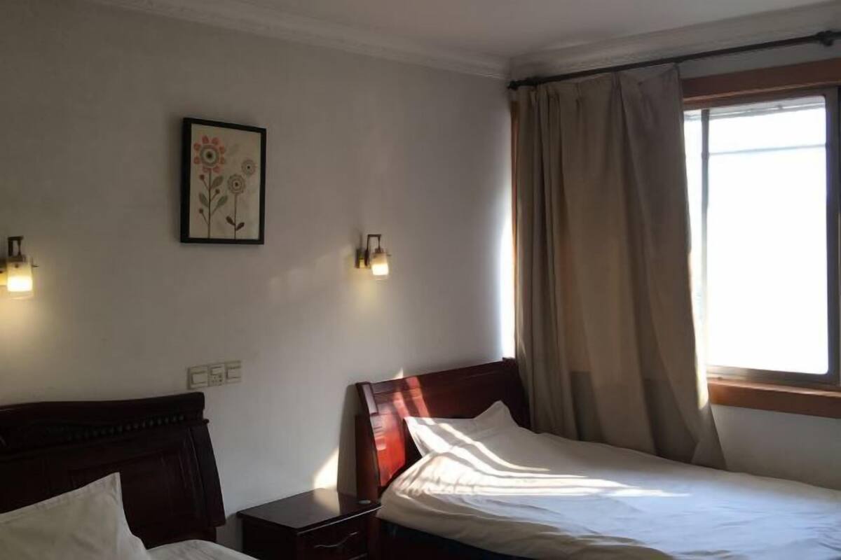 A single bed with a wooden headboard, a bedside table, a wall-mounted light, and a window with curtains.