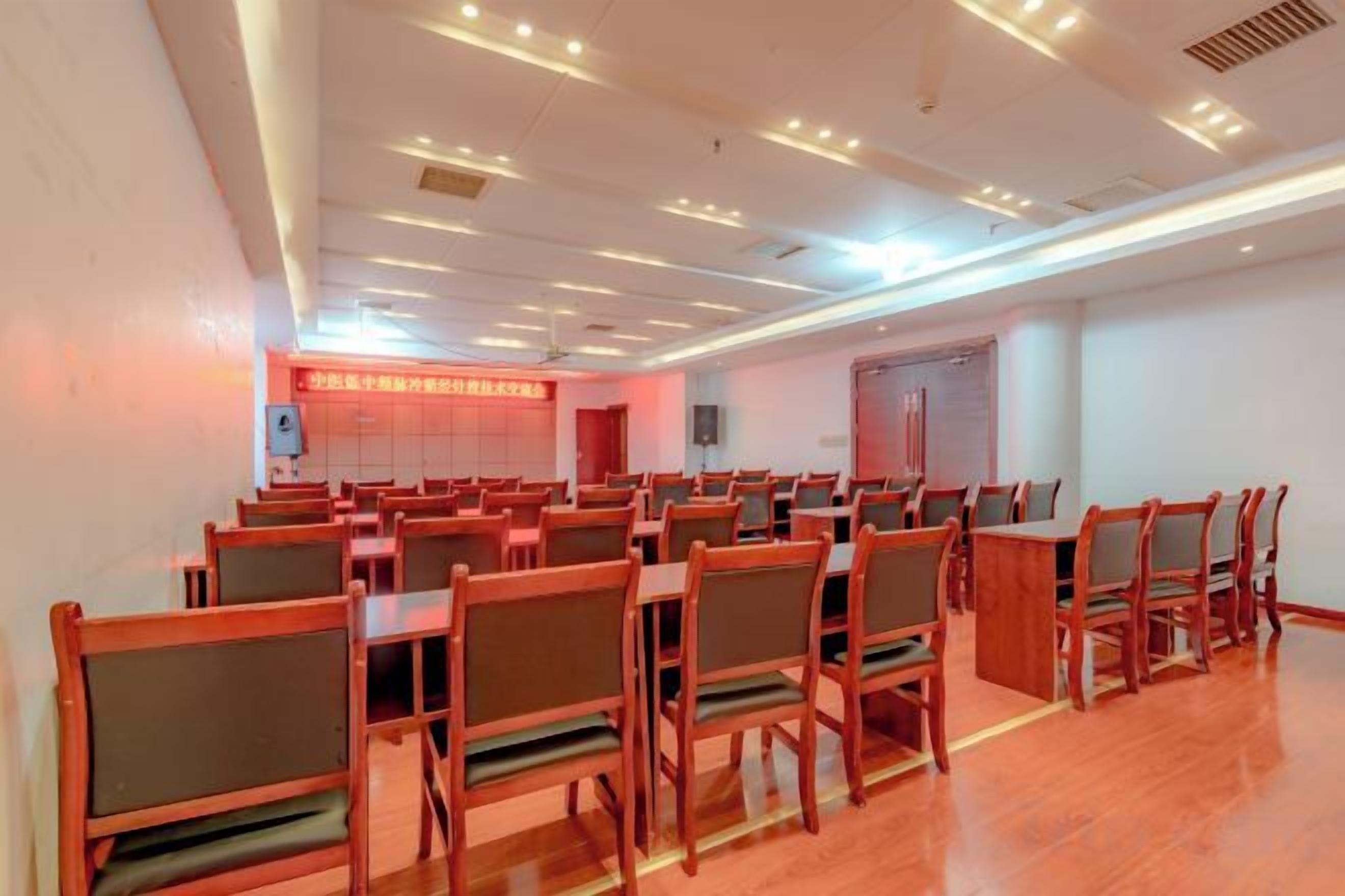 Un salón de conferencias con filas de sillas y mesas de madera, un cartel rojo en la pared y una puerta al fondo.