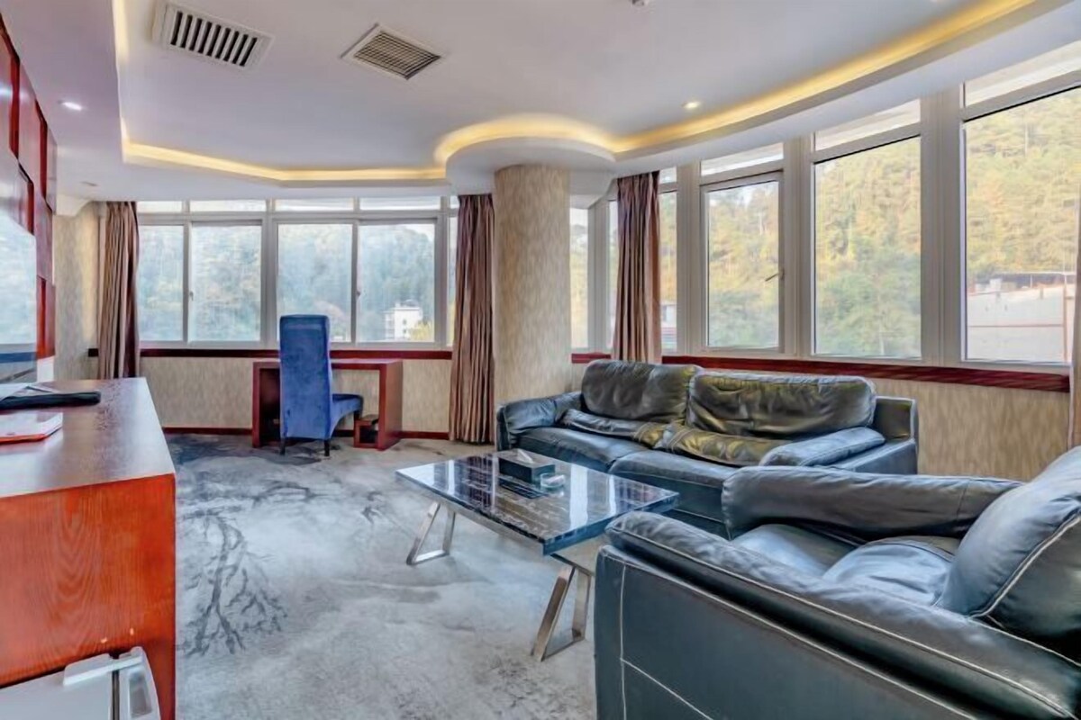 Une chambre d’hôtel moderne, dotée d’un canapé en cuir, d’une table basse en verre, d’un bureau en bois et de grandes fenêtres offrant une vue sur l’extérieur.
