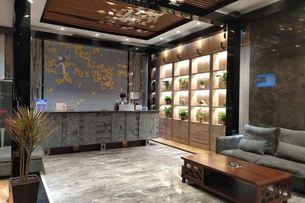 Zona de recepción con mostrador de mármol, una recepcionista, un sofá, una mesa de centro y una pared con estanterías que albergan plantas.