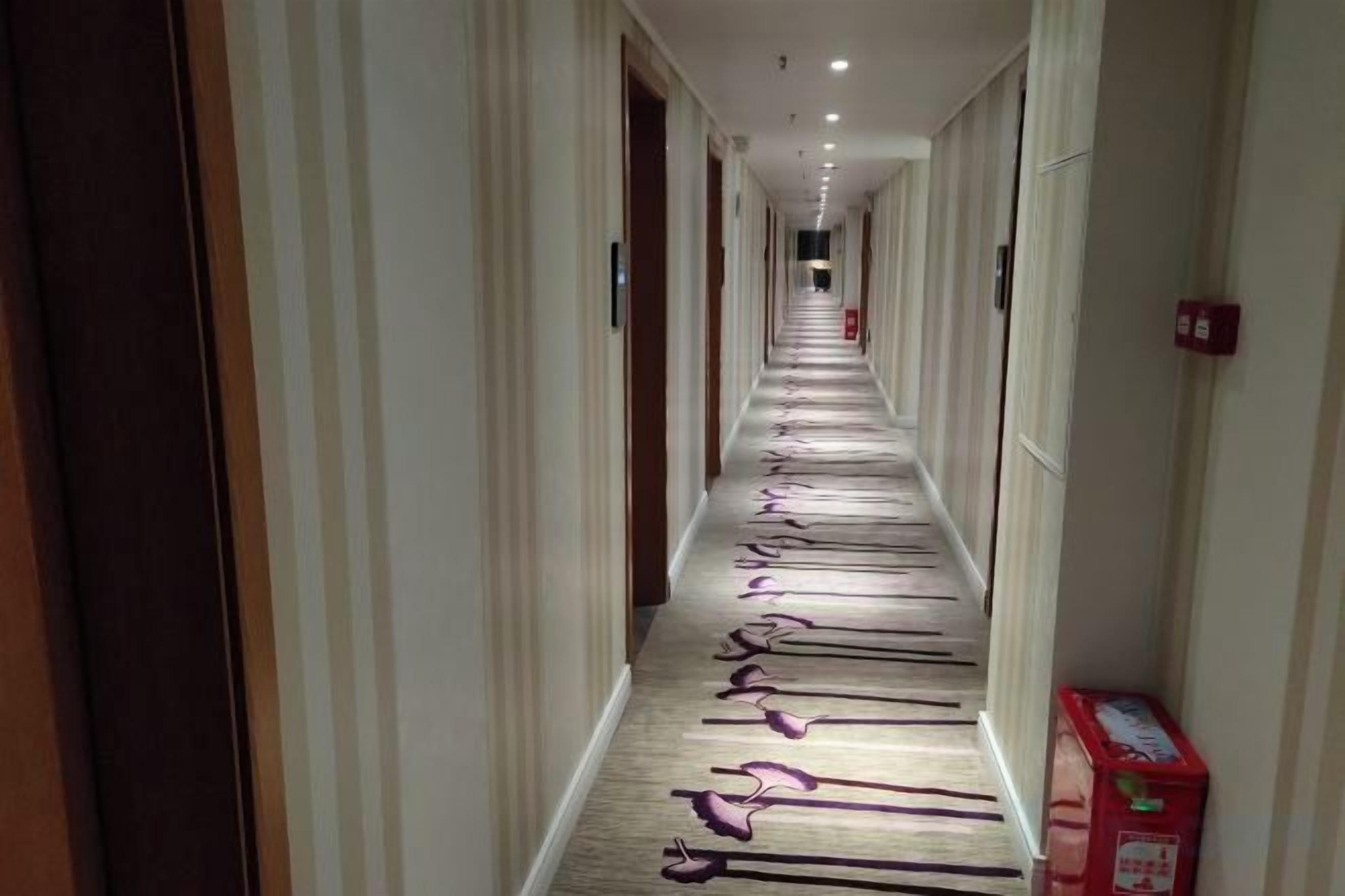 Un pasillo de hotel con puertas a ambos lados, piso cubierto de alfombra y un extintor en la pared derecha.