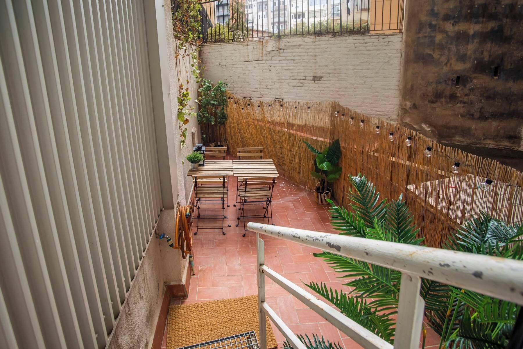 A narrow outdoor patio with a wooden table and chairs, surrounded by a bamboo fence and potted plants.