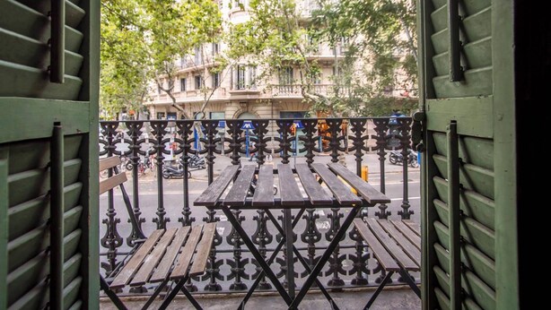 Une porte ouverte donne sur une cour intérieure où se trouvent une table en bois et des bancs, le tout entouré de rampes en métal noir ouvragé.