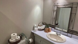 Baño con inodoro, lavabo y espejo. Hay artículos de aseo en el mostrador y una planta sobre el lavabo.