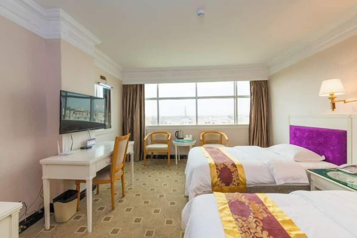 Una habitación de hotel con dos camas, un escritorio con televisor, una ventana con cortinas y una lámpara.